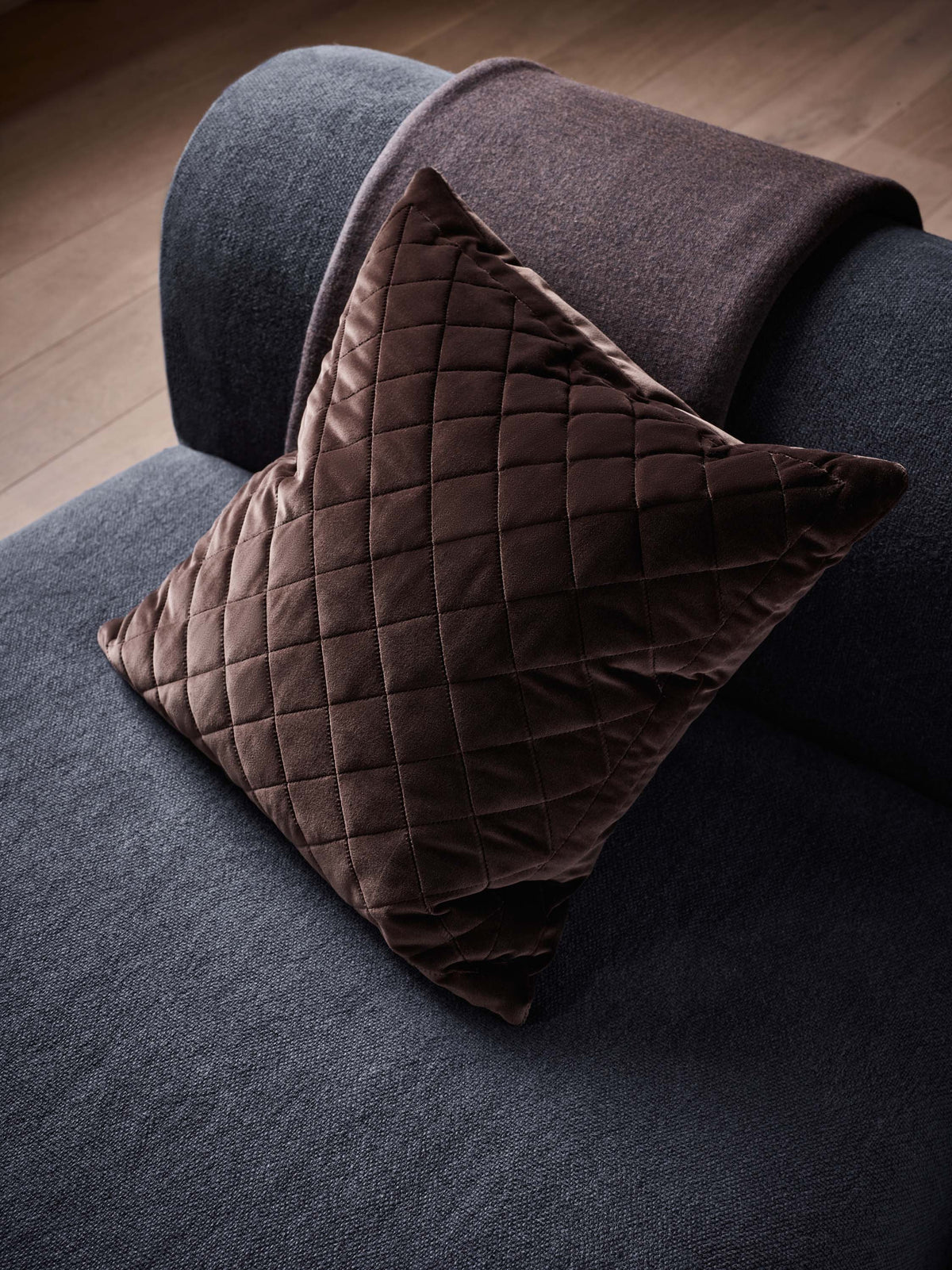 A dark brown quilted velvet throw pillow resting on a dark gray sofa with a matching brown throw blanket draped over the arm, set against a wooden floor background.