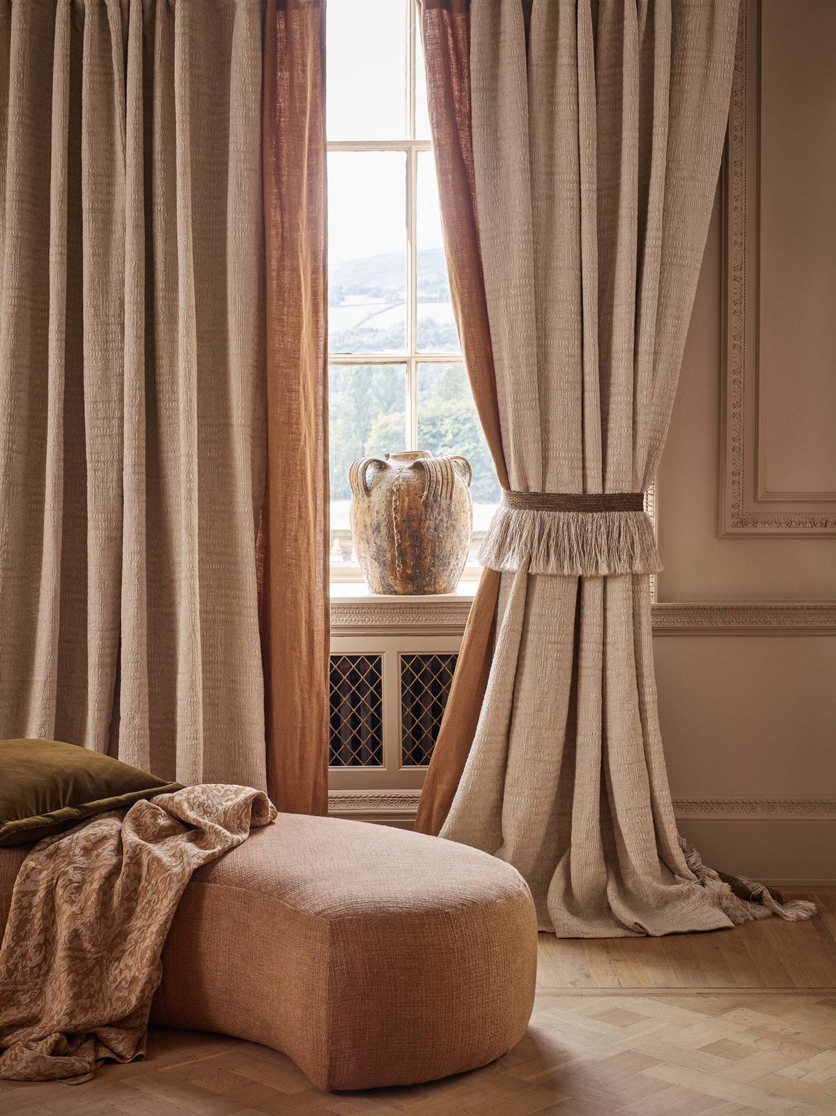 A styled interior scene showing floor-length curtains in warm beige and tan tones hanging around a window, with a matching upholstered ottoman in the foreground and a vase on the windowsill.