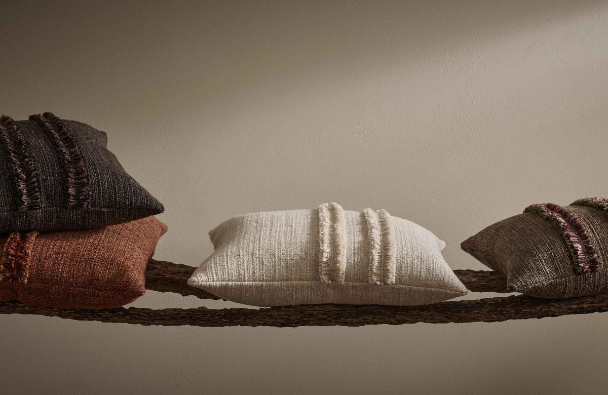 Four textured pillows in brown, red, white, and green on a wooden shelf against a beige wall.